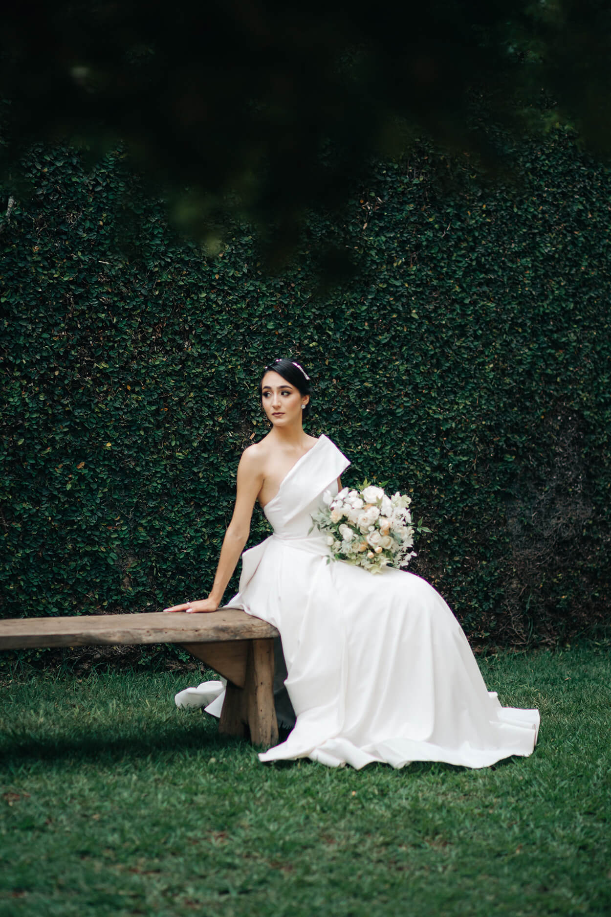 noiva sentada em banco de madeira ao ar livre segurando buquê redondo com flores brancas e vestido com ombro único