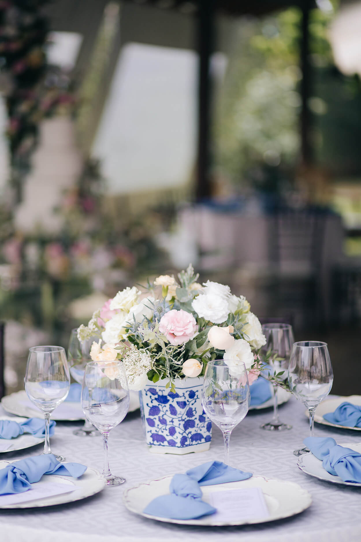 mesa redonda com toalha branca e mesa posta azul com vasos de flores brancas e rosas no centro