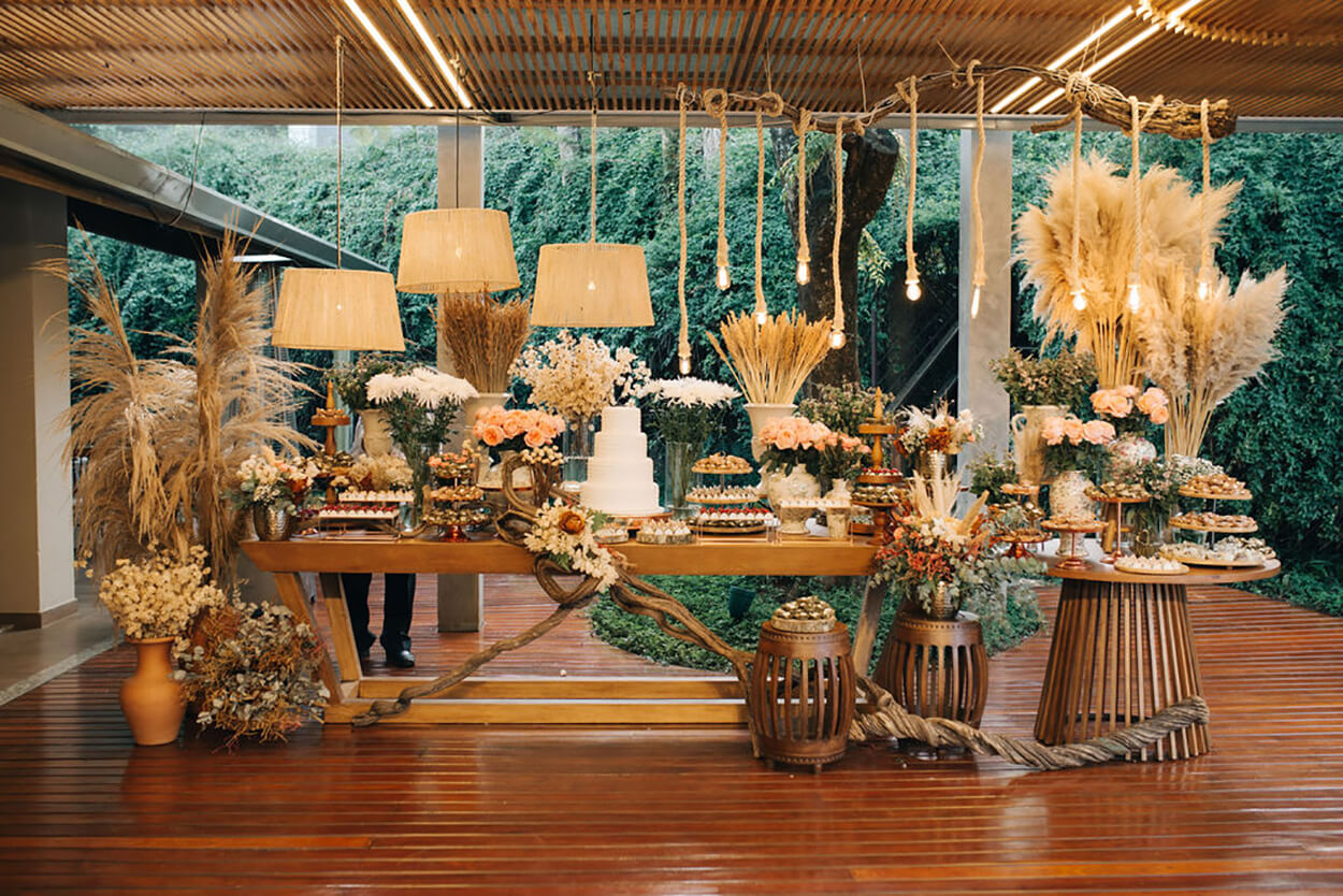 mesa de madeira com bolo de casamento com 4 andares e arranjos com flores e capim dos pampas e luminárias rústicas com corda