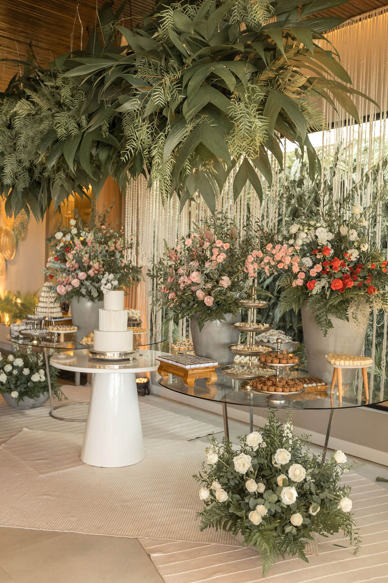 salão com duas mesas de vidro com bandejas de doces e vasos com flores brancas rosas e vermelhas e mesa branca redonda no centro com bolo de casamento