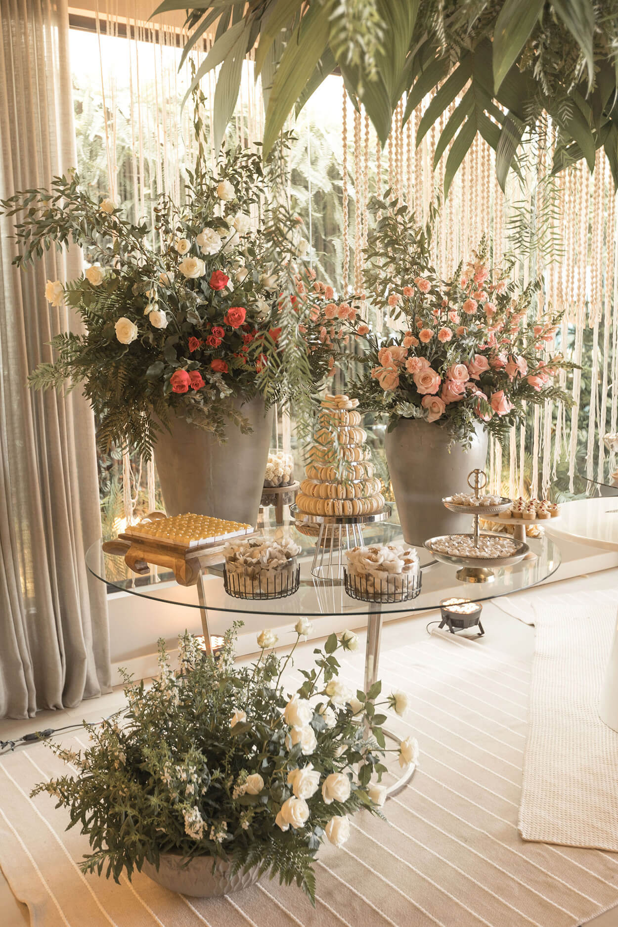Casamento estilo moderno e Mesa de vidro com torre de macarrons brancos e bandejas com doces e dois grandes vasos cinzas com flores vermelhas rosas e brancas