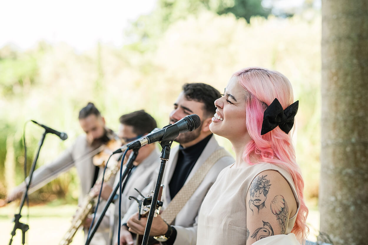 mulher com cabelo rosa e homens cantando nos microfones ao ar livre