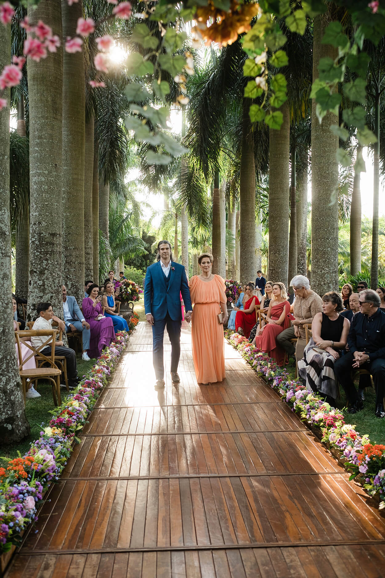 entrada do noivo com terno azul de mãos dadas com a mãe com vetsido laranja claro