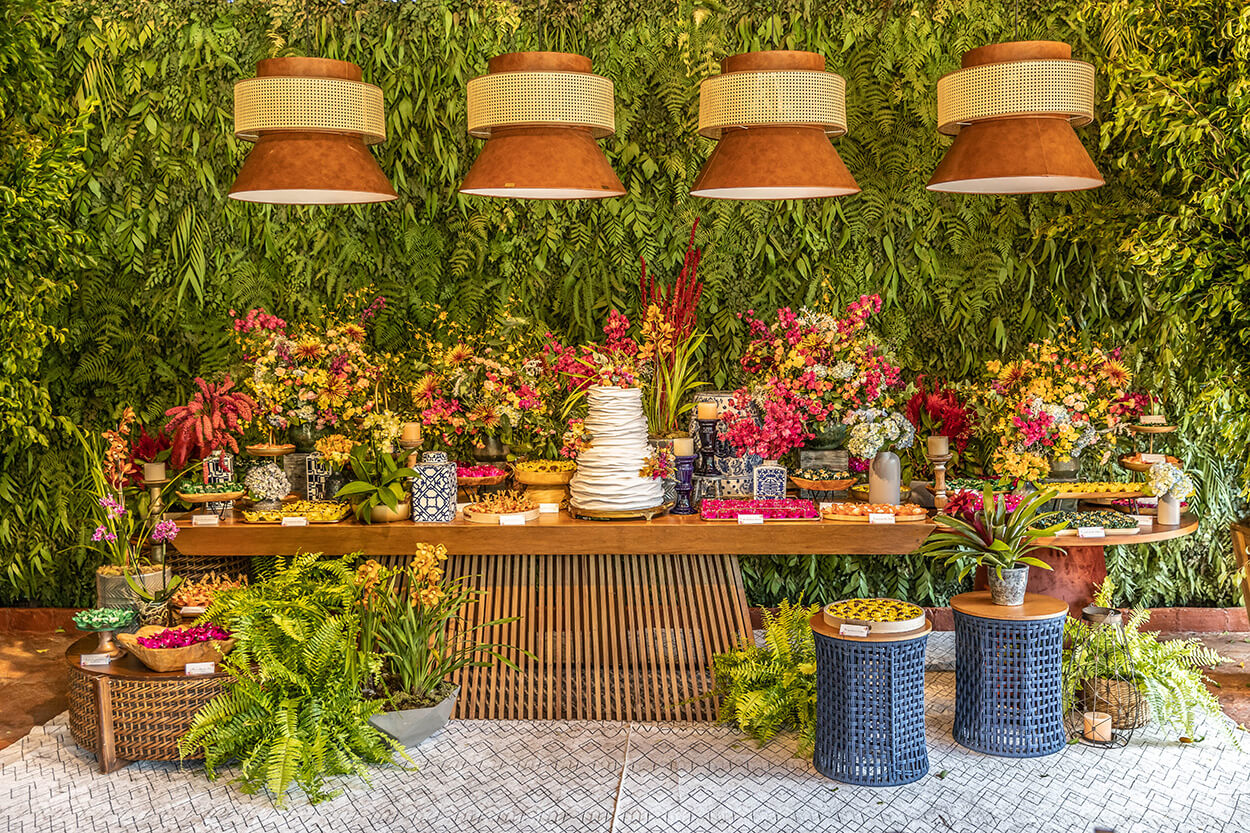 salão com painel com plantas e mesa de madeira com bolo de casamento e vasos com flores rosas e amarelas