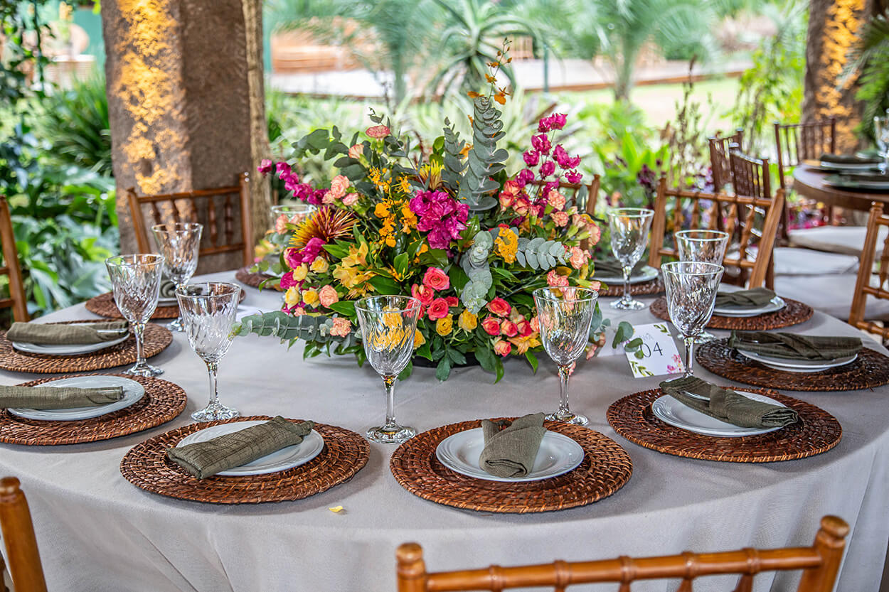 mesa redonda com toalha branca posta com sousplat de palha com flores amarelas e rosas no centro