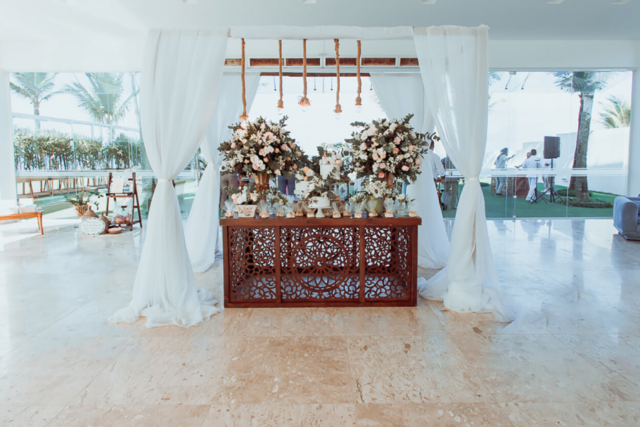 salão com mesa de madeira com vaos de flores brancas e rosas bolo de casamento e doces