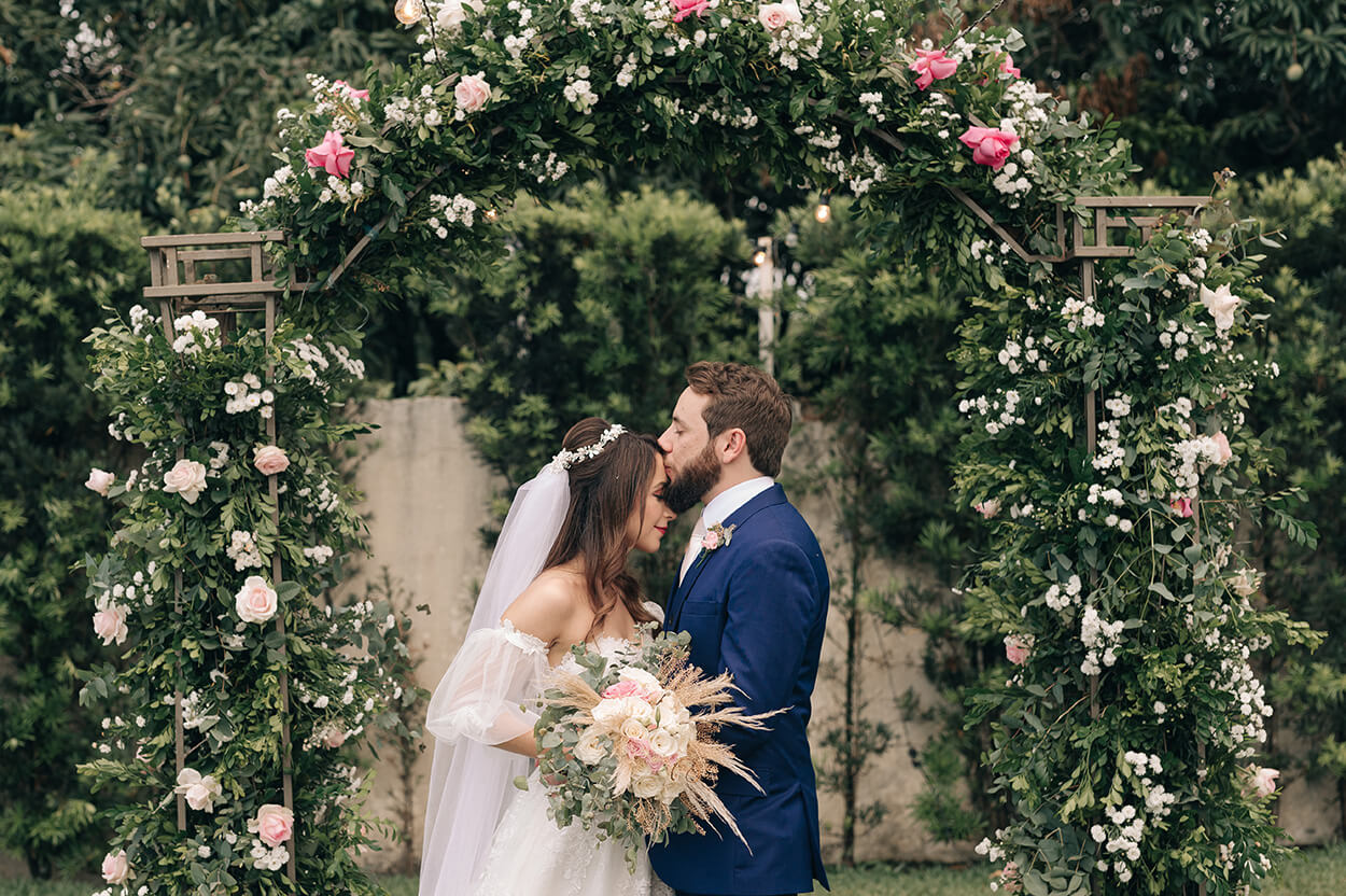 noivo beijando a testa da noiva com arco com rosas brancas e rosas