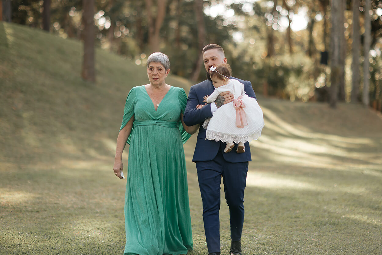 entrada do noivo com terno azul segurando filha bebê com vestido branco e mãe com vestido verde 