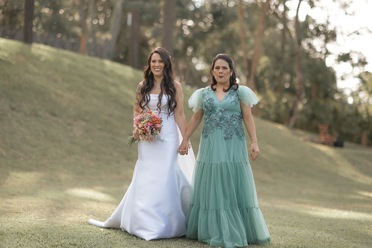 noiva com vetsido semi sereia segurando buquê colorido de mãos dadas com a mãe com vestido verde com mangas bufantes e bordados florais