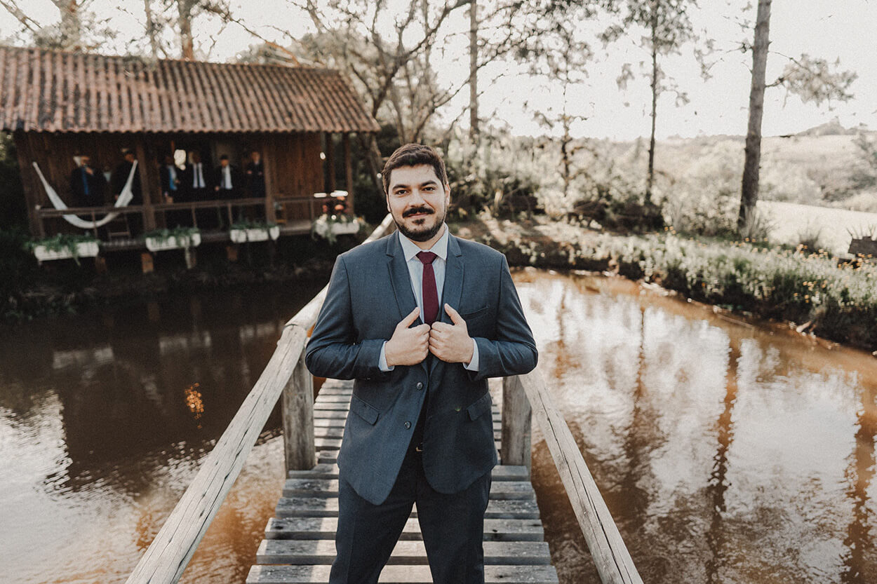 noivo com terno azul com colete e gravata vermelha no meio d aponte de madeira