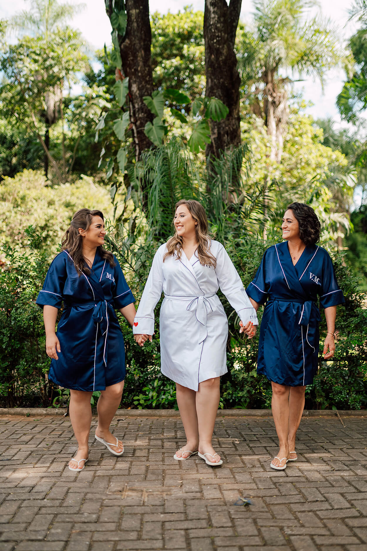 noiva com robe branco de mãos dadas com duas mulheres com robes azuis escuros