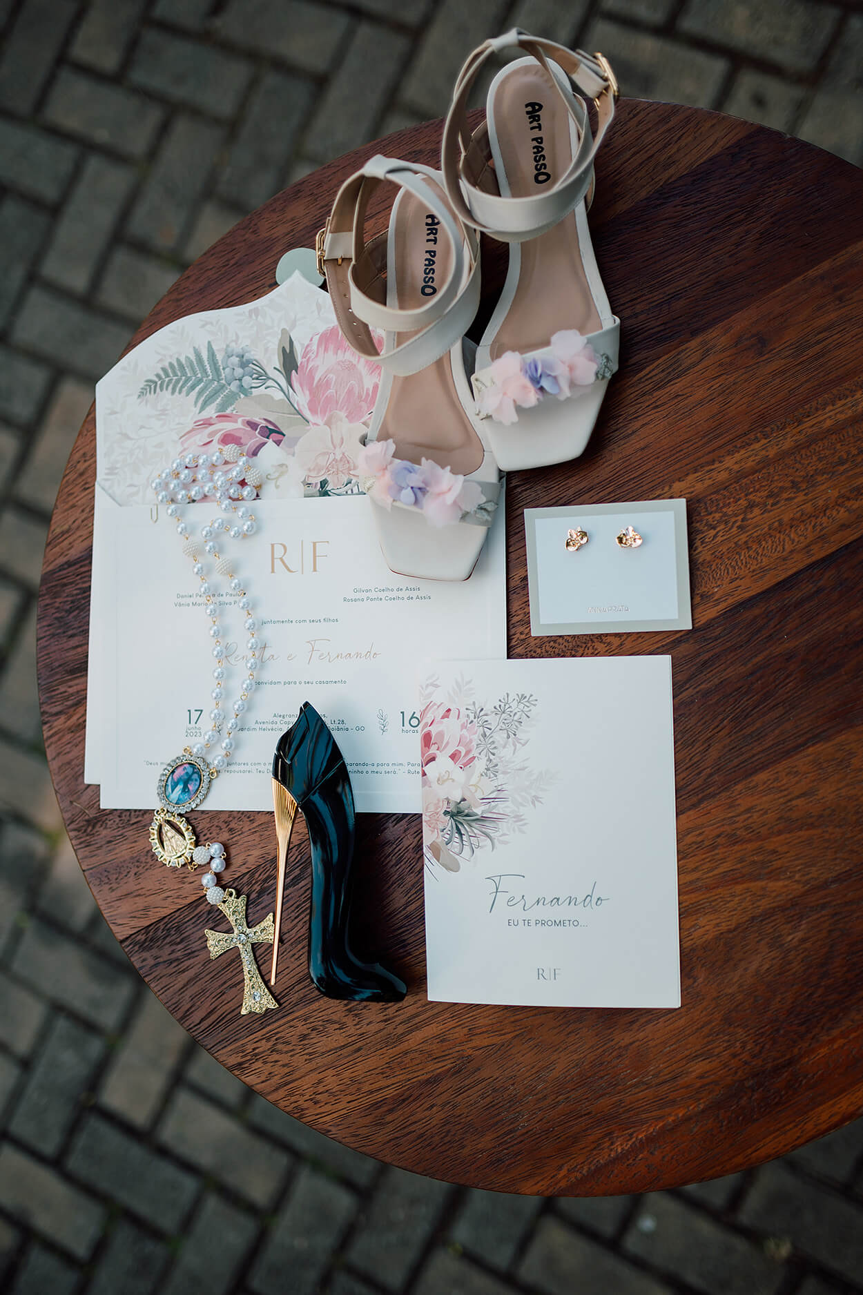convite de casamento branco com estampa de flores ao lado de sandála branca com flores coloridas e perfume em forma de salto alto e corrente com cruz