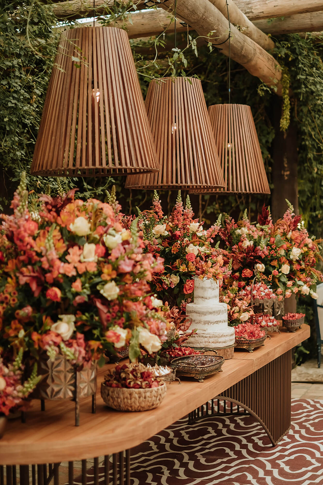 salão rústico com mesa de madeira com bolo de casamento semi espatulado e arranjos com flores terracota e bandeja com doces e três luminárias rusticas