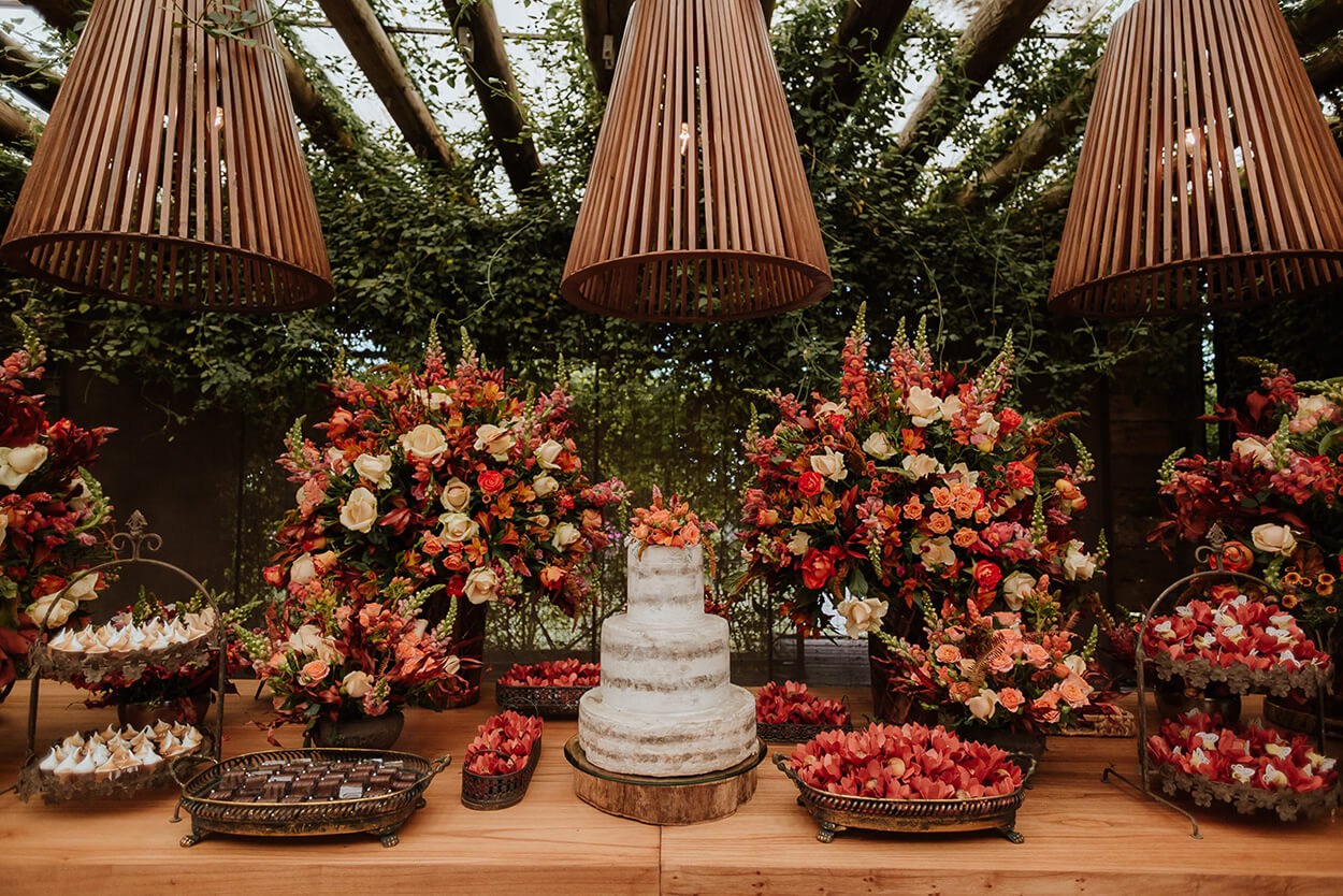 salão rústico com mesa de madeira com bolo de casamento semi espatulado e arranjos com flores terracota e bandeja com doces