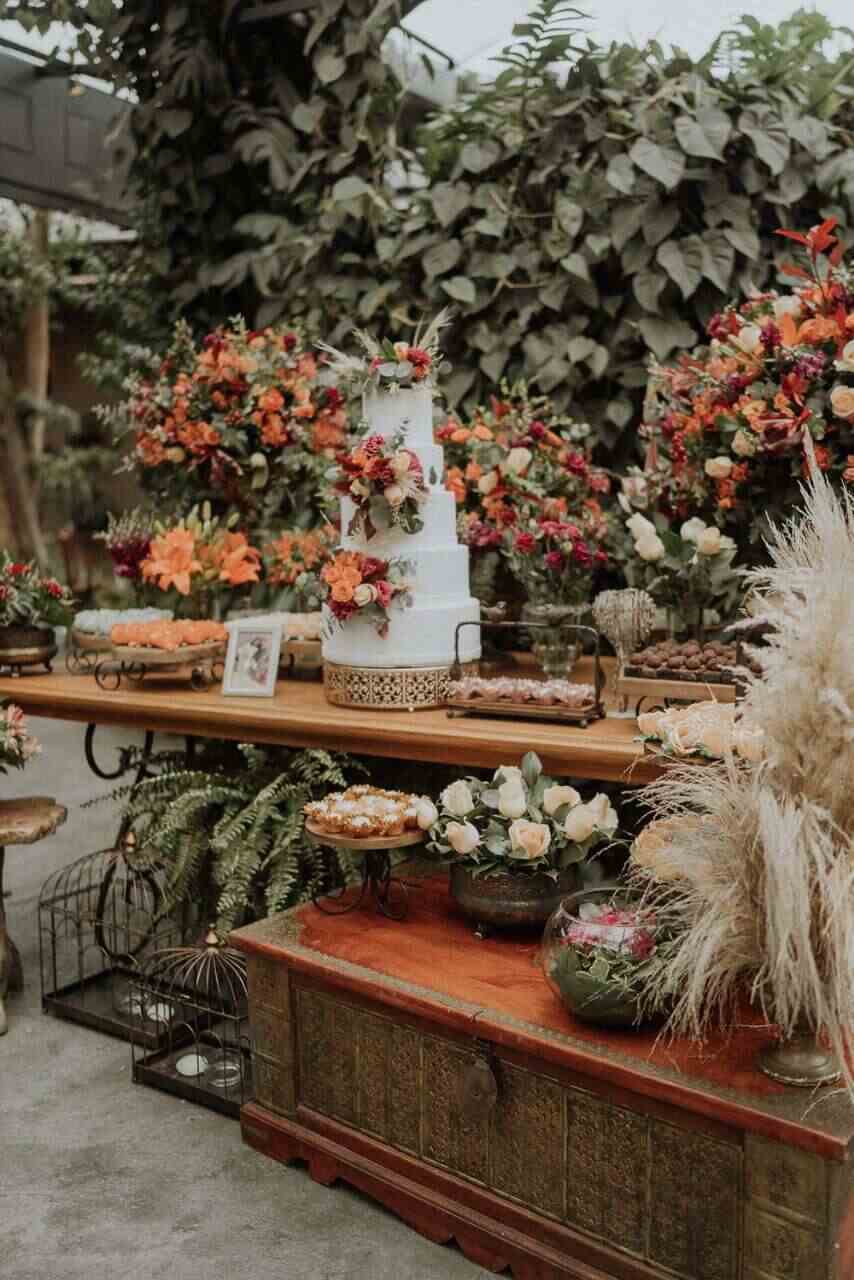 mesas de madeira com bandejas com doces bolo de casamento de três andares com flores laranjas e vermelhas e no chão gaiolas de ferro retrô com samambaias