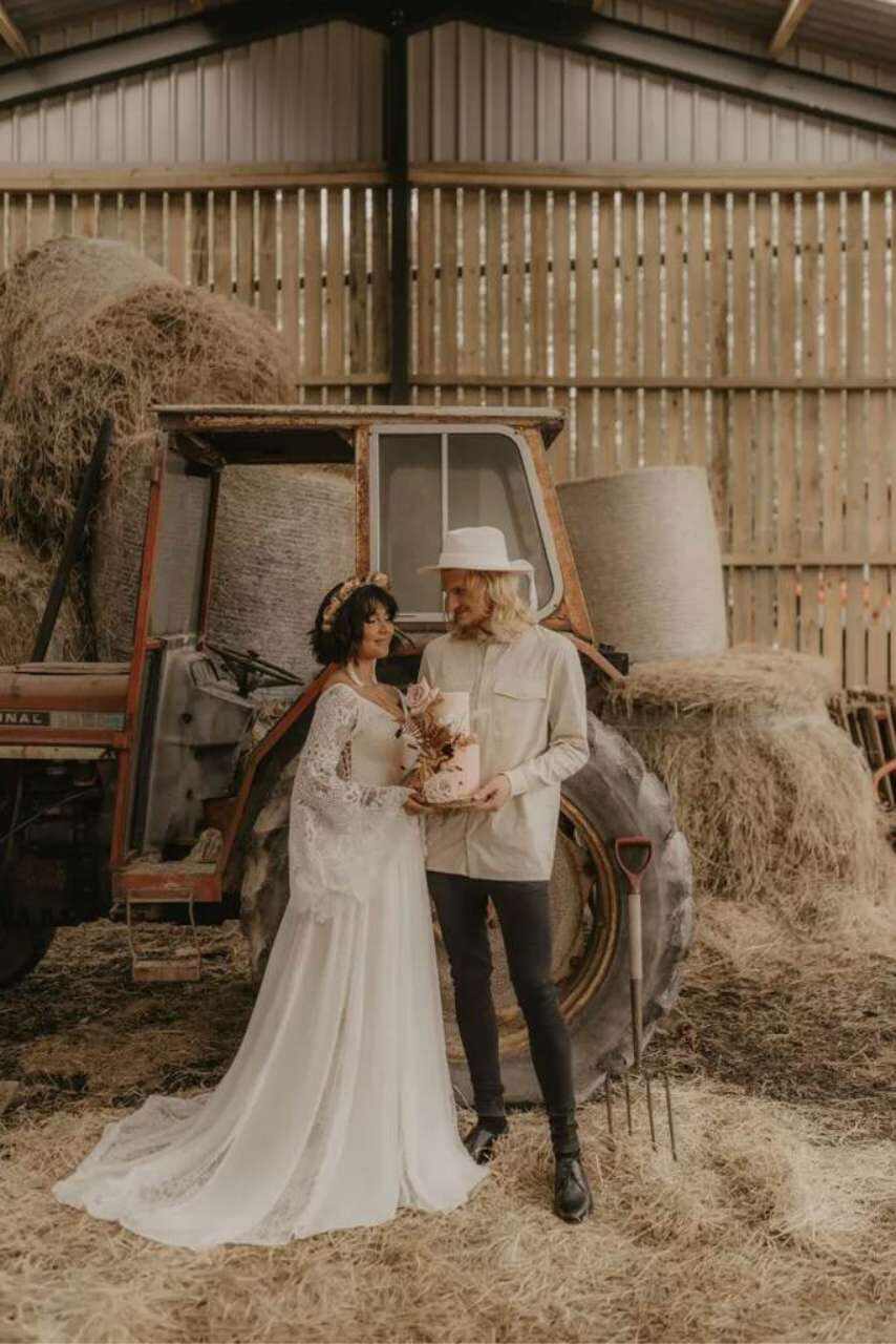 noiva com vestido com manga longa ao lado do noivo com chapéu seguranod bolo dentro do celeiro