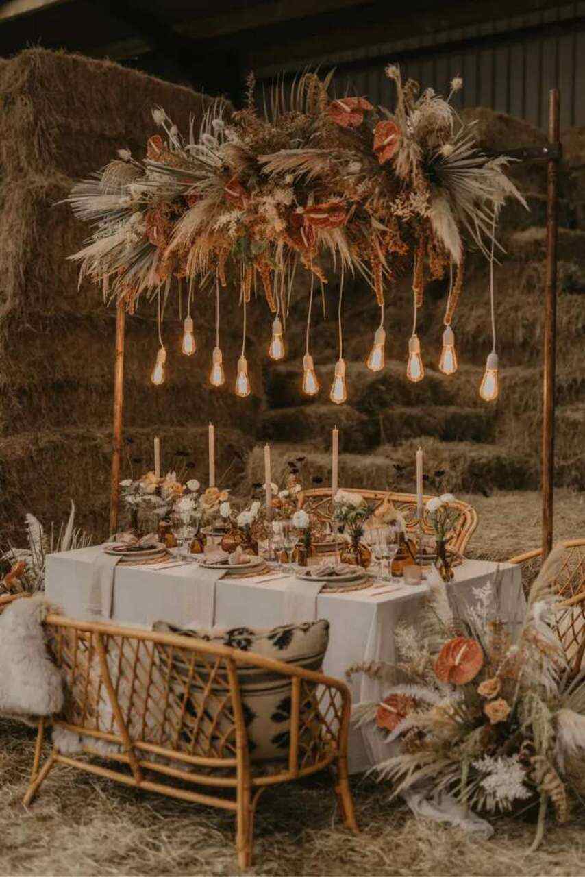mesa posta com velas flores e arranjos pendentes com flores laranjas capim dos pampas e lâmpadas bancos rústicos em meio ao celeiro