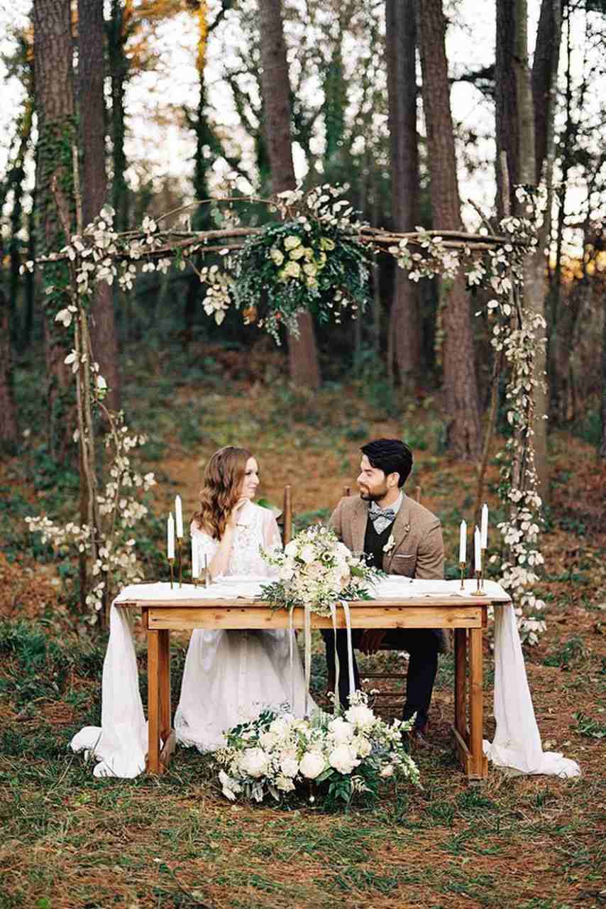 noiva e noivo sentados a mesa no meio da floresta decorada com tecido branco e flores brancas