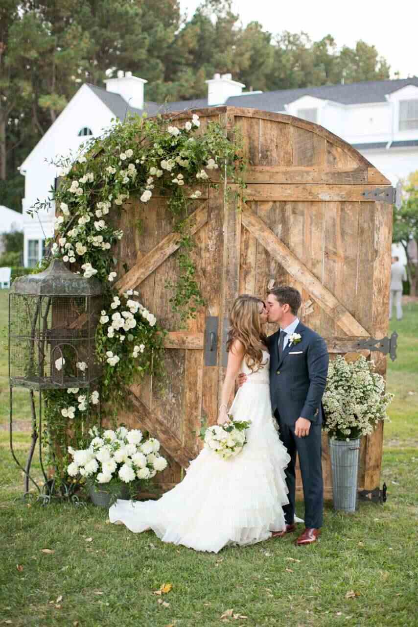 noivos se beijando na frente de portão de madeira antigo decorado com folhagens e flores brancas