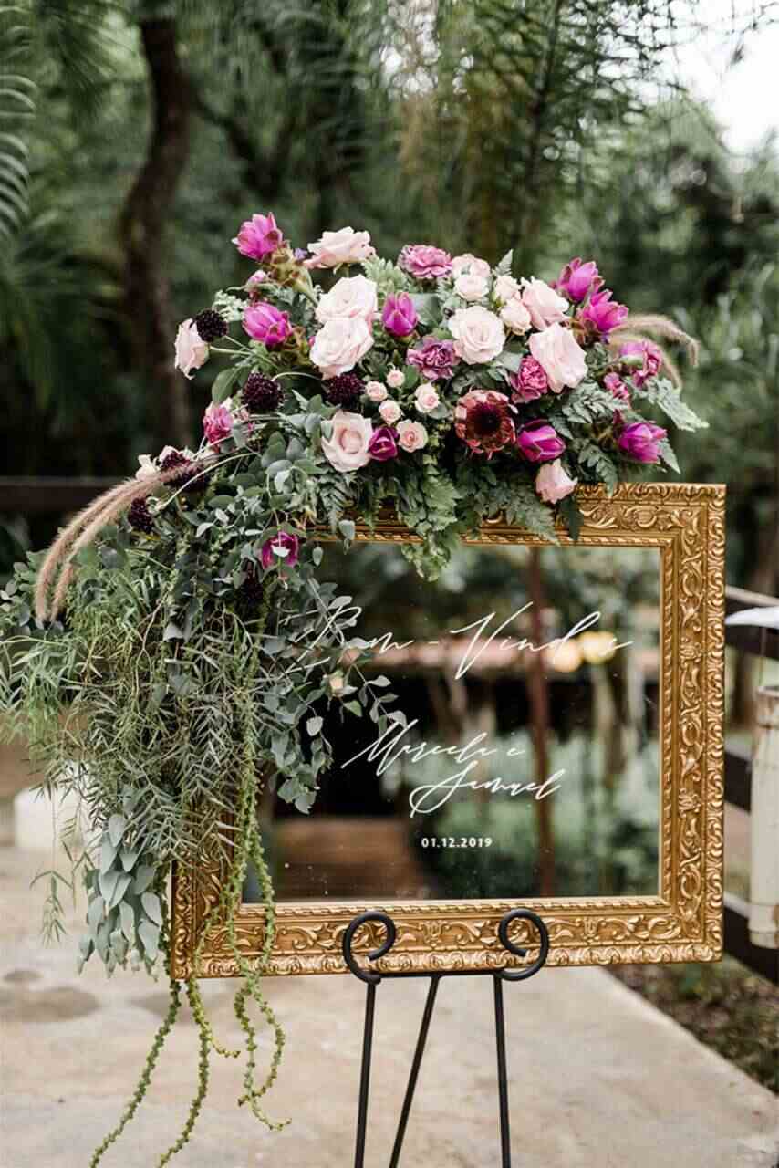 espelho com o nome dos noivos e moldura dourada antiga com o arranjo em cima com flores brancas e rosas