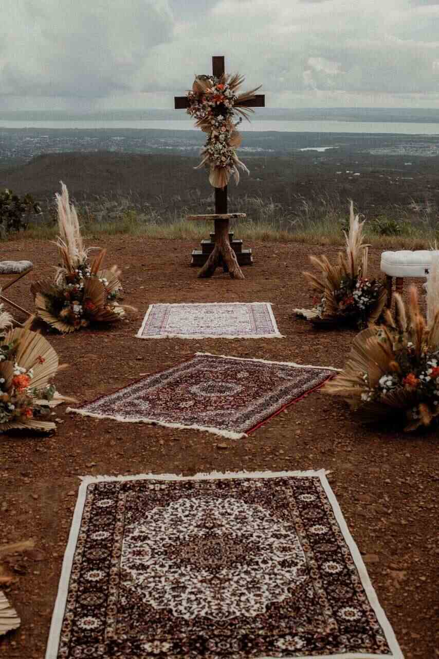altar com cruz de madeira com folhas secas e capim dos pampas na terra com vista para as montanhas e três tapetes étnicos