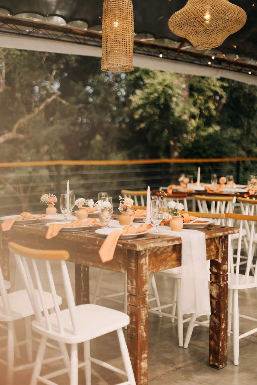 mesas de madeira rústica com cadeiras brancas e mesa posta com guardanapos laranjas