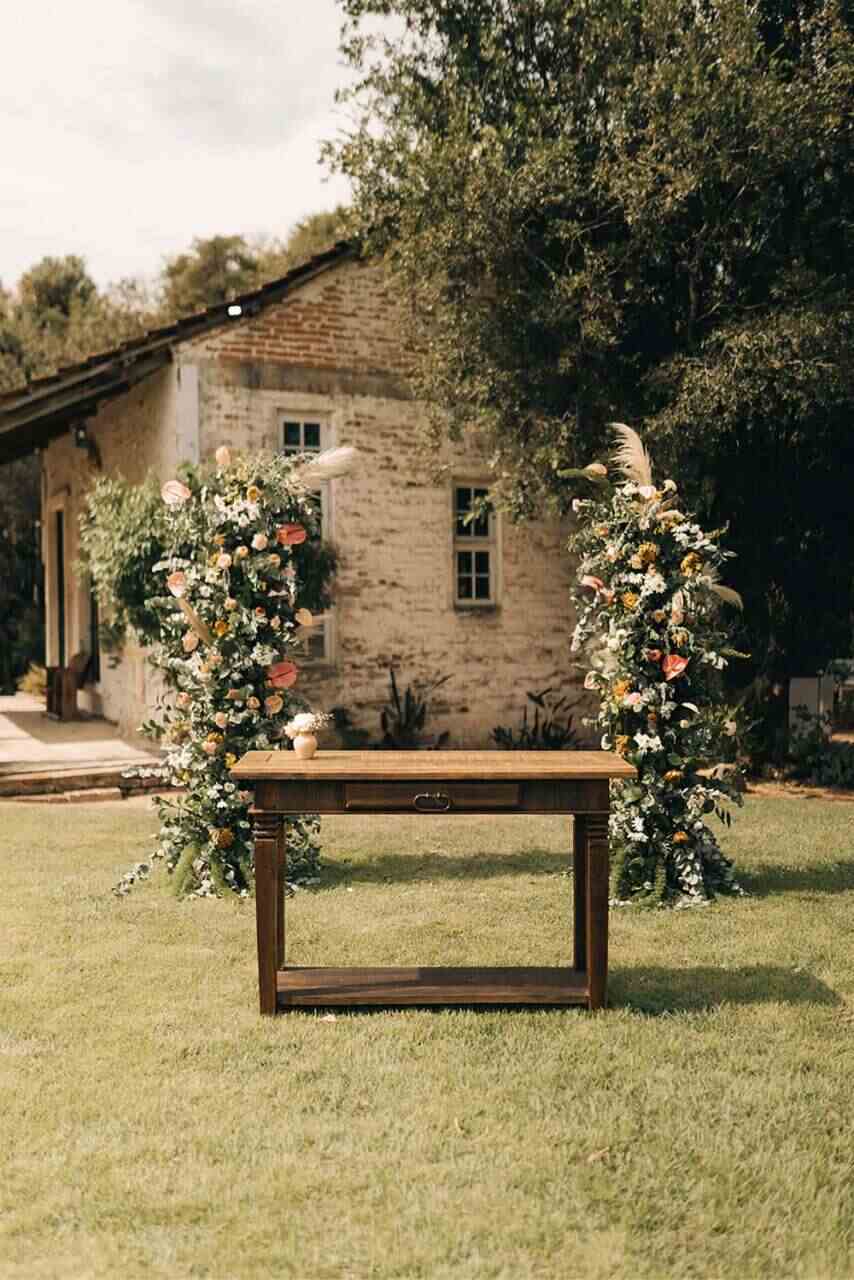 mesa de madeira escura rústica com altar com flores laranjas brancas e vermelhas e capim dos pampas ao fundo casa antiga com tijolos a mostra