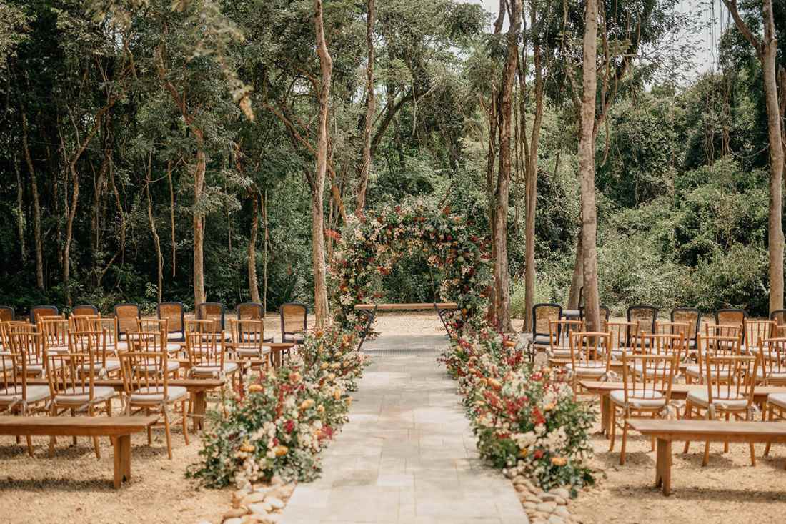 bancos e cadeiras de madeira claras de frente par a floresta e corredor da noiva todo decorado com flores brancas amarelas e vermelhas e arco floral para cerimônia