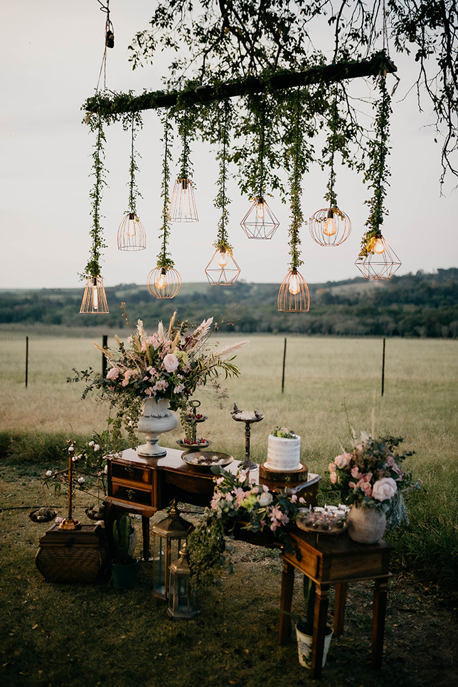 mesas de madeira com bolo de casamento e flores co de rosa e arranjo com plantas pendentes e lâmpadas para decoração de casamento rústico