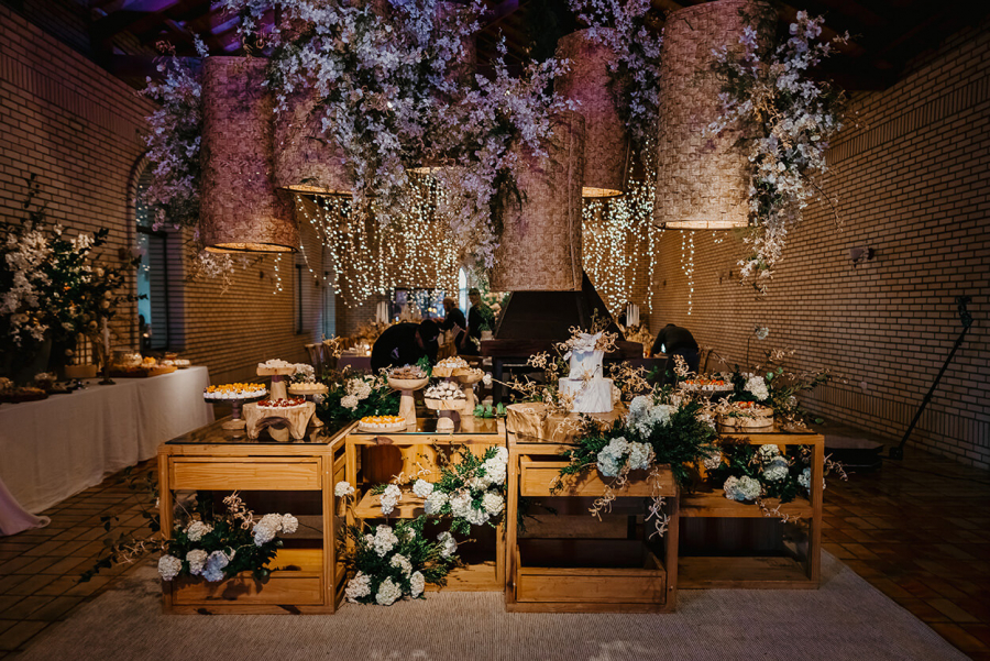 mesa de madeira com bolo de casamento branco e bandejas de madeira com doces e acima dela luminarias grandes e flores pendentes