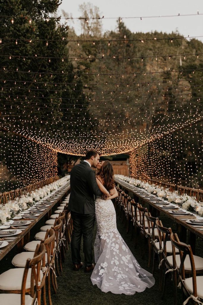 noivos de costas se beijando no corredor entre as mesas postas cobertas com varal de luzes