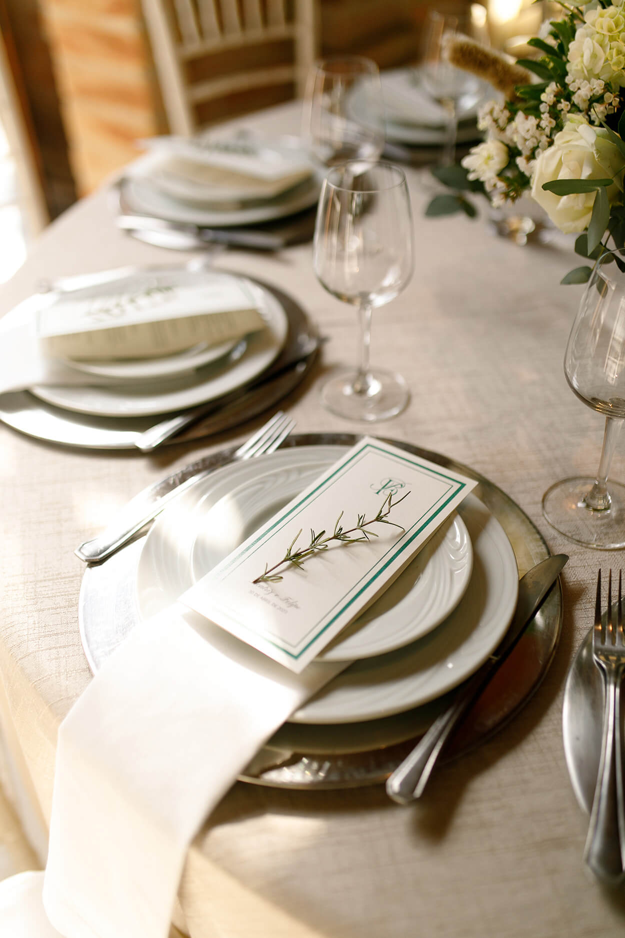 mesa com toalha branca posta com menu sobre o prato branco com ramo de alecrim