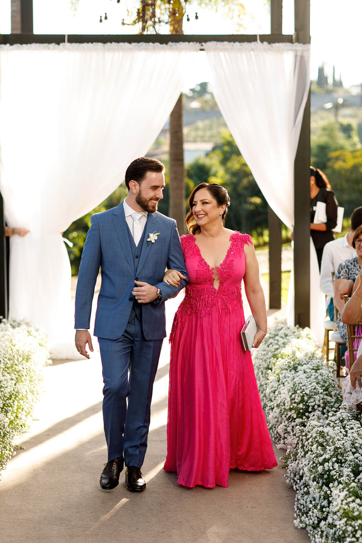 entrada do noivo com terno azul e gravata branca ao lado da mãe com vestido longo e pink