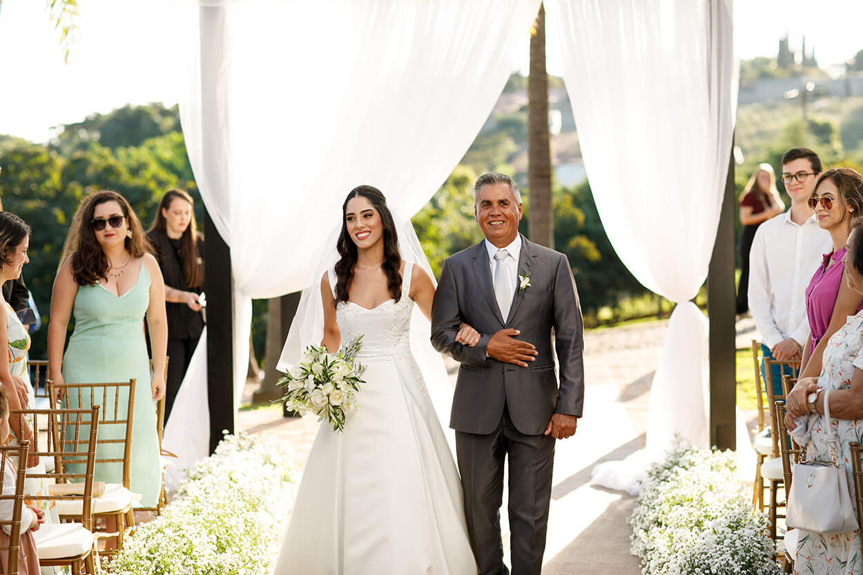 entrada da noiva com vestido minimalista e elegante com buquê com rosas brancas a lado do pai com terno cinza e gravata branca