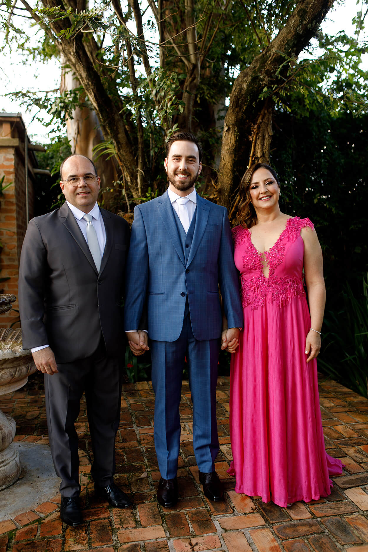 noivo com terno e colete azul riscado com gravata branca de mãos dadas com o pai com terno preto e gravata branca e mãe com vestido longo pink 
