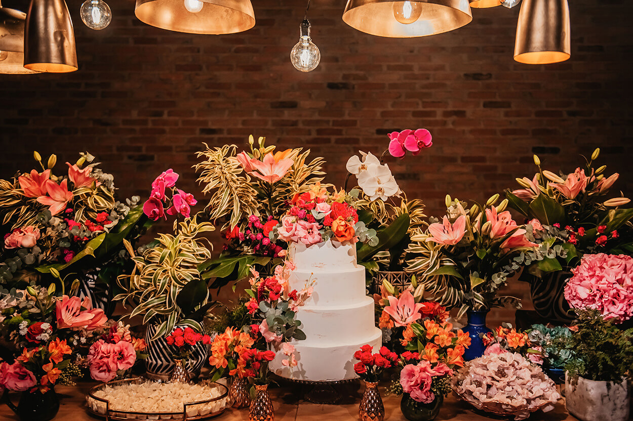 mesa de madeira com bolo de casamento com quatro andares com flores no topo e vasos com flores rosas ao lado