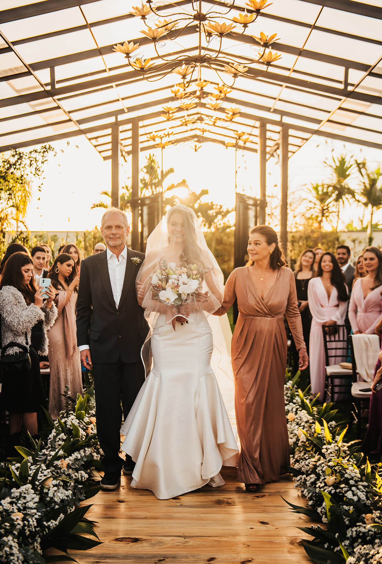 entrada da noiva com véu no rosto acompanhada do pai com terno preto e mãe com vestido rose com decote v