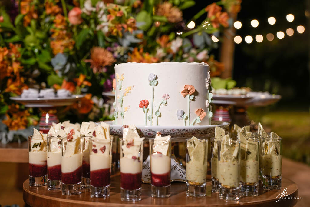 bolo de casamento branco com floes espatuladas coloridas e em volta copinhos com doces