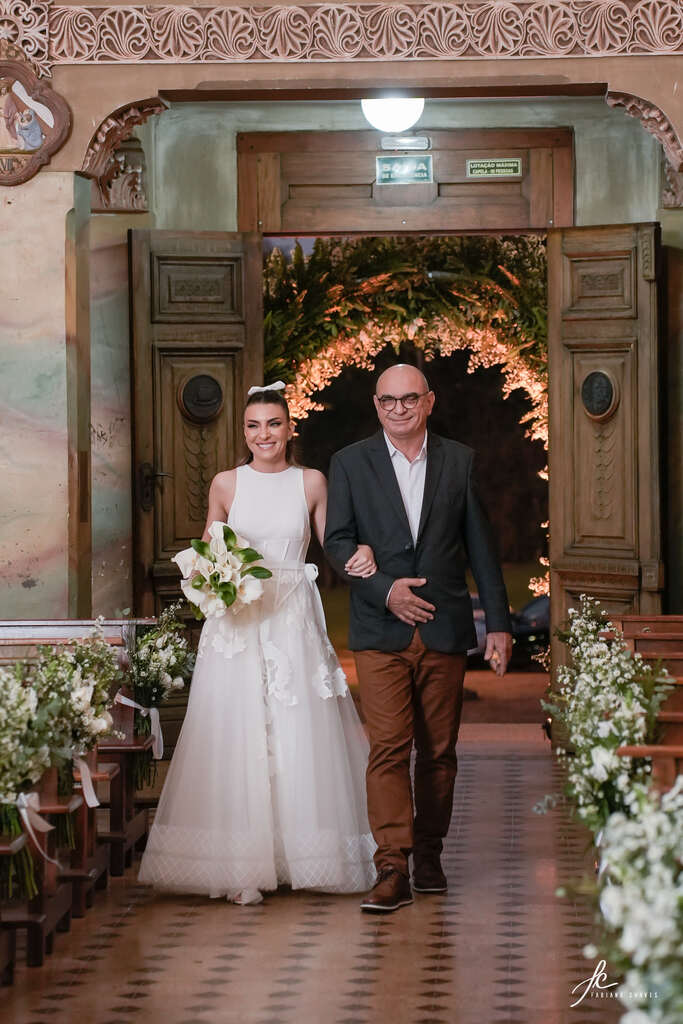 entrada da noiva com vetsido minimalista segurando buquê com flores brancas ao lado do pai com terno cinza escuro e calça marrom