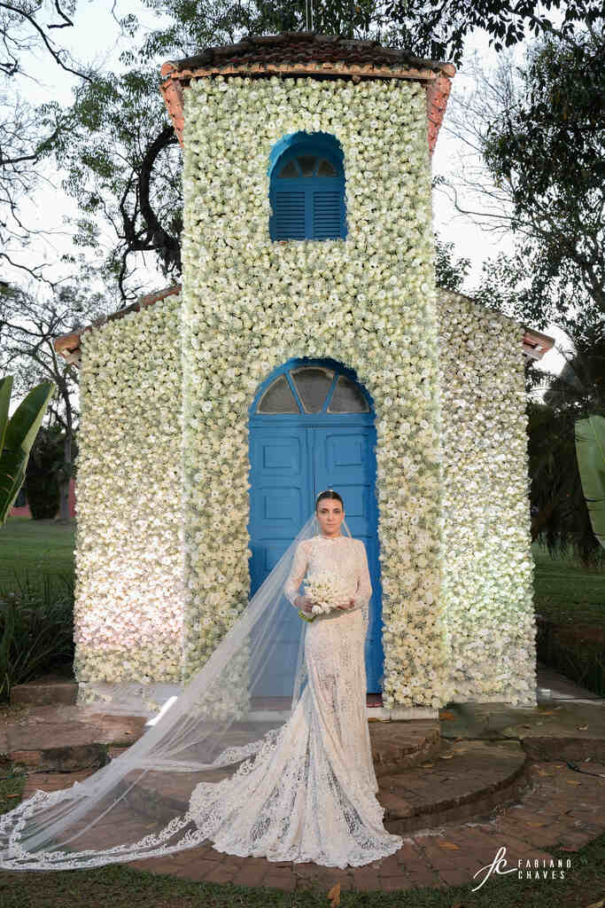 mulher com vestido de noiva longo com mangas segurando buquê com tulipas e mantilha na frente de capela decorada com flores brancas