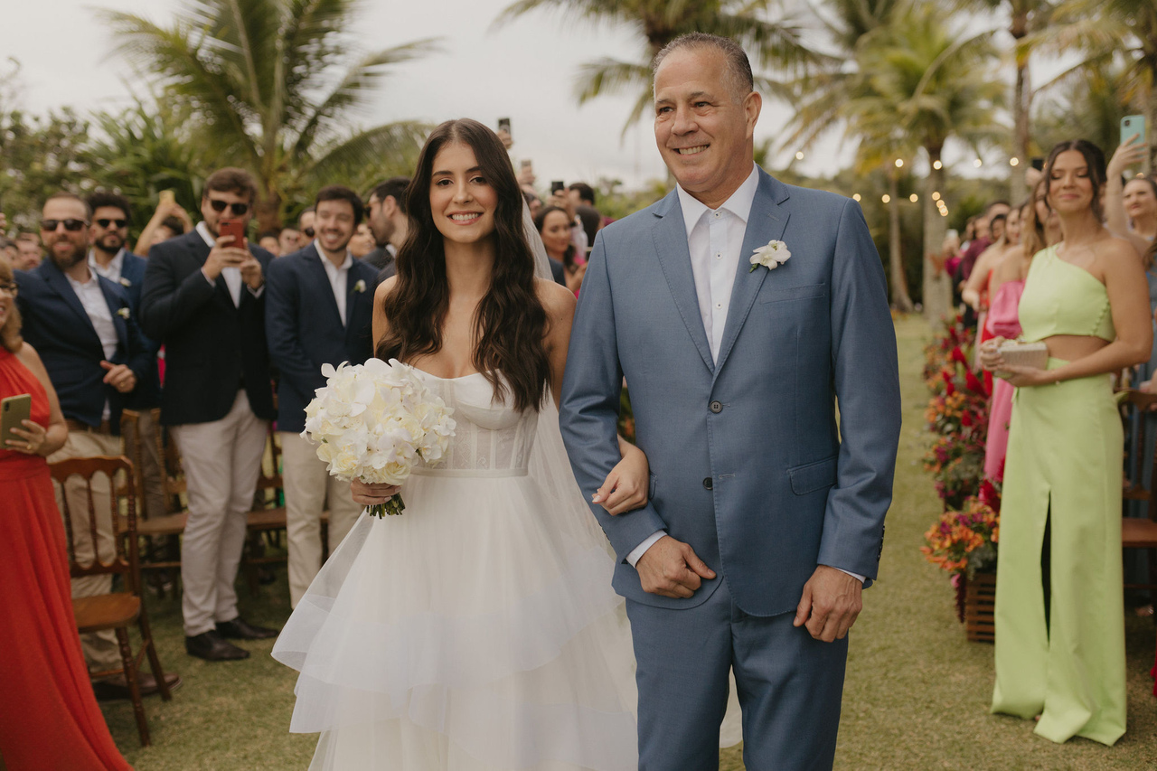 entrada da noiva com buquê com flores brancas ao lado do pai com terno azul com flor branca na lapela