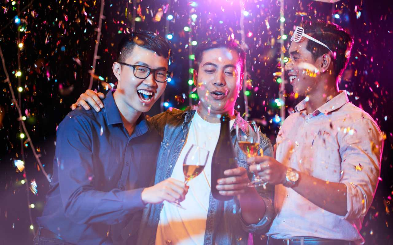 três homens asiáticos brindando com champagne em despedida de solteiro de noite com confete metalizado caindo