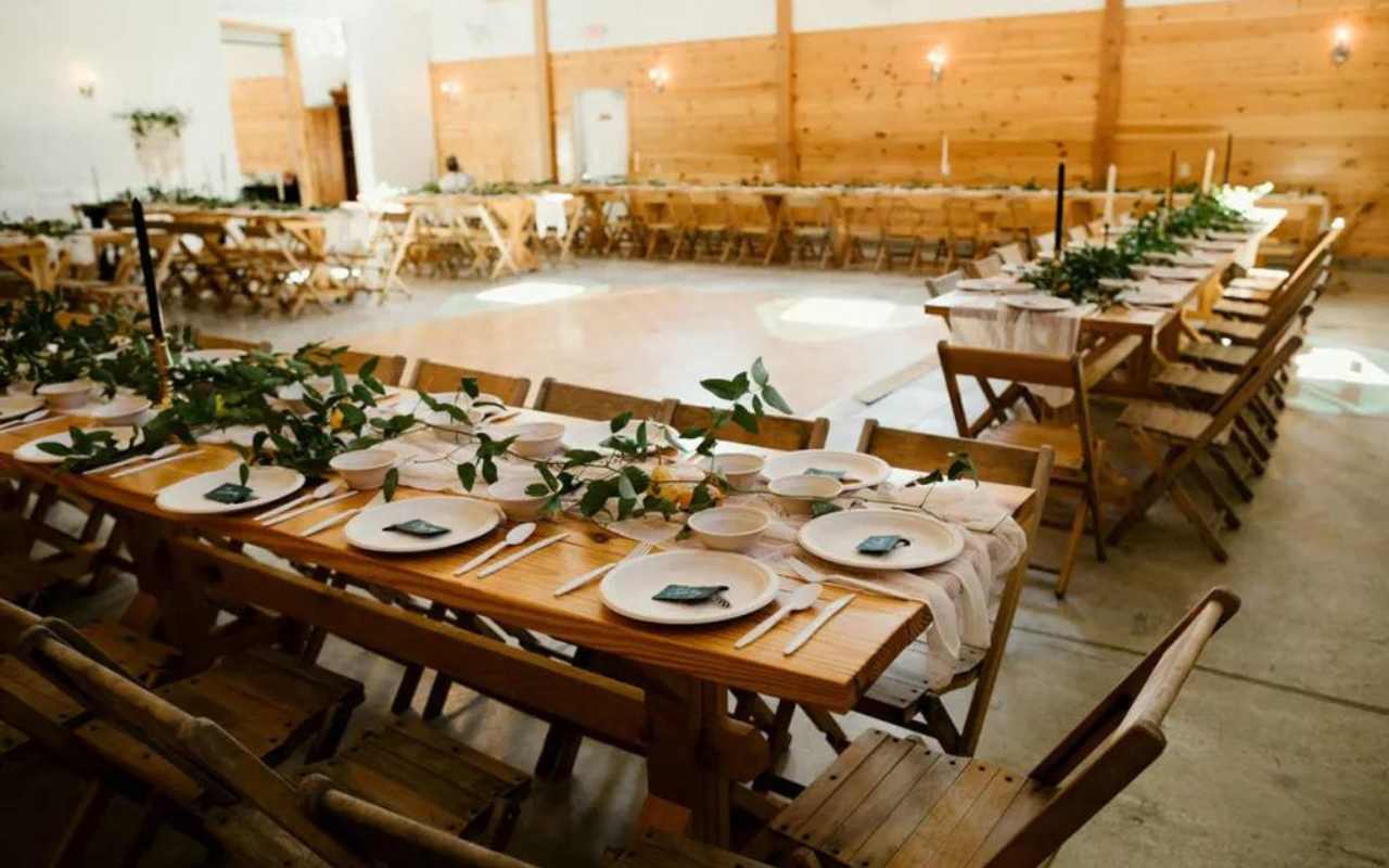 recepção de casamento em salão interno com decoração rústica, mesas de madeira, folhagens de eucalipto, velas, castiçais, louças branca e frutas