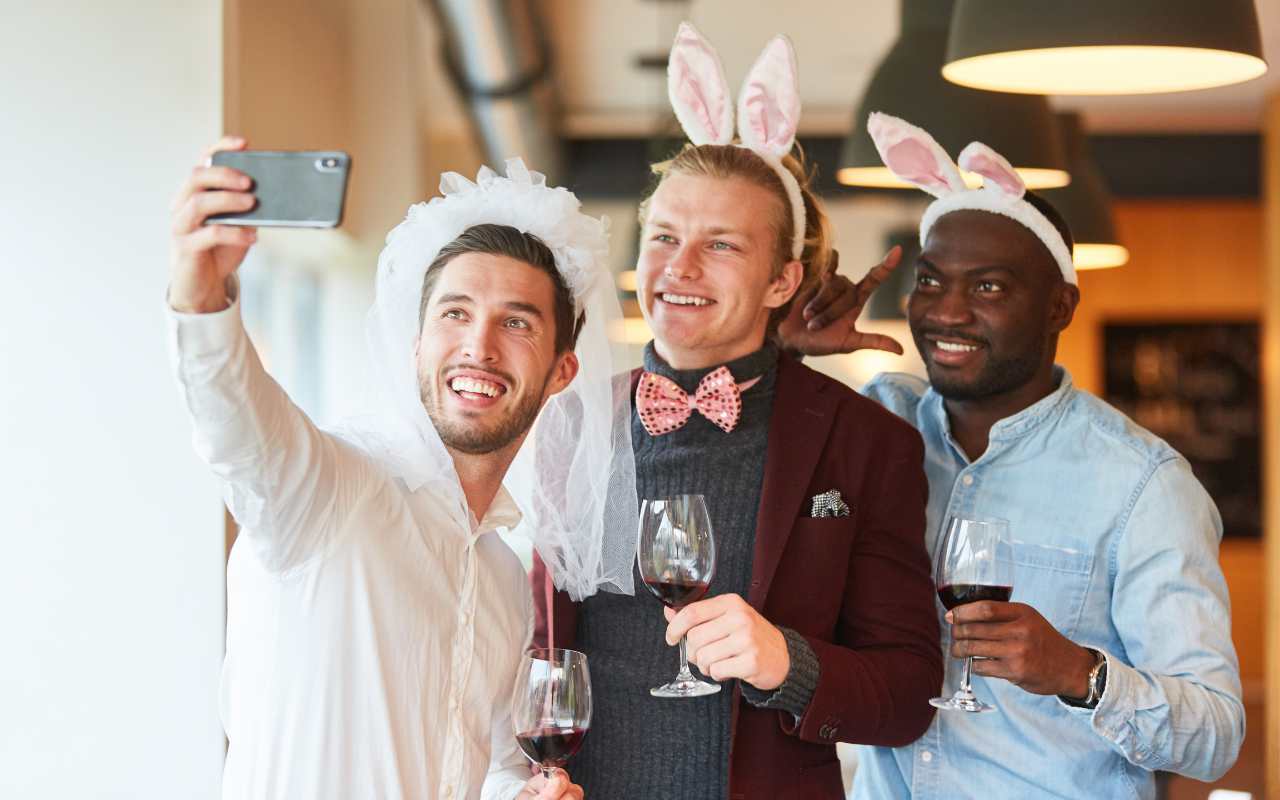 três homens tirando selfie divertida em despedida de solteiro com copos de vinho na mão e acessórios na cabeça