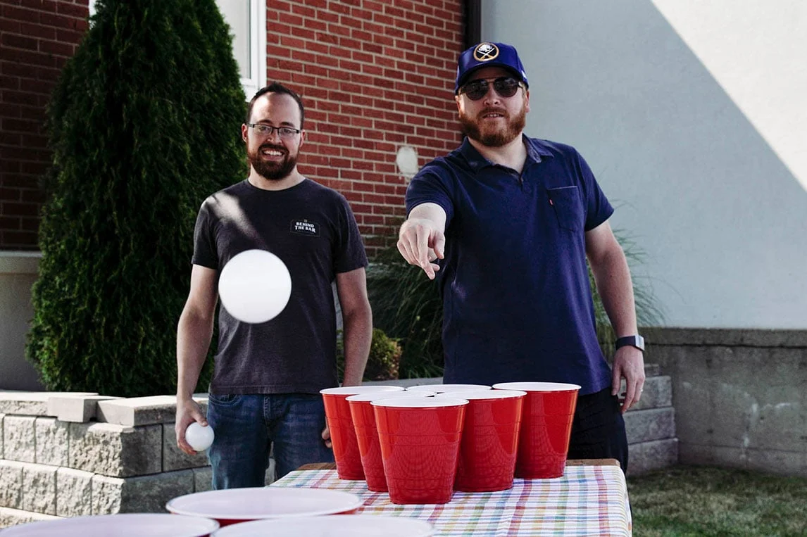 dois homens arremessando bolinha de ping pong em copos vermelhos durante jogo de beers pong em despedida de solteiro