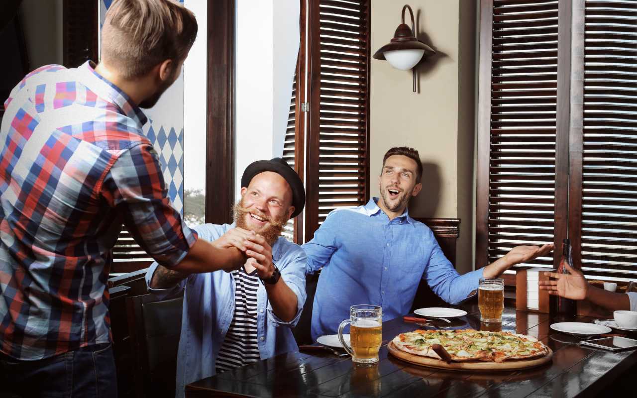 homens comendo pizza em restaurante. Um deles está cumprimentando outro que acaba de chegar