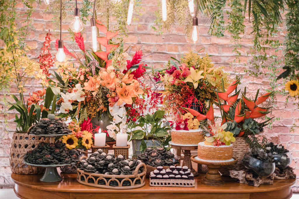 mesa de madeira com bandejas com doces e vasos com orquídeas brancas, girassois, lírios amarelos e laranja e outras flores e dois bolos de rolo