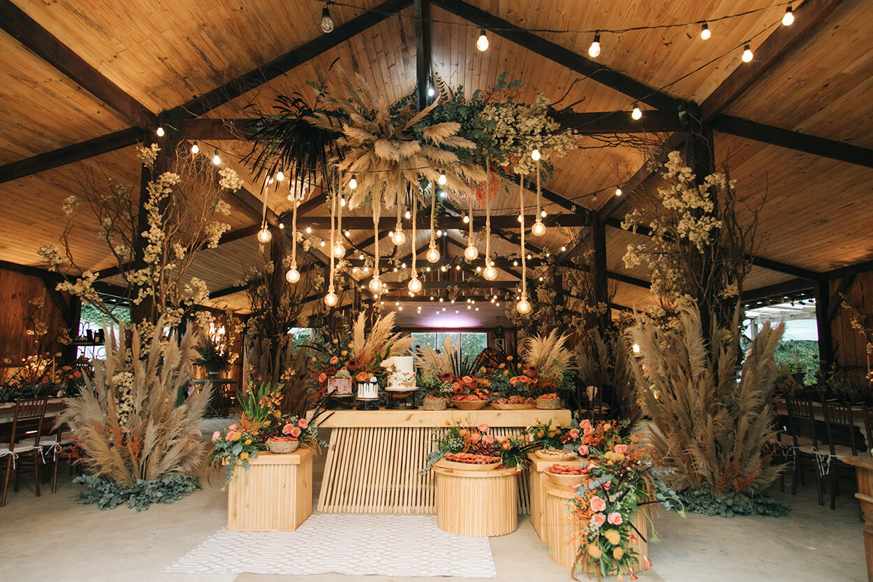 mesas de madeira com bolo de casmaento e vasos com flores e enfeites com capim dos pampas dentro do salão rústico