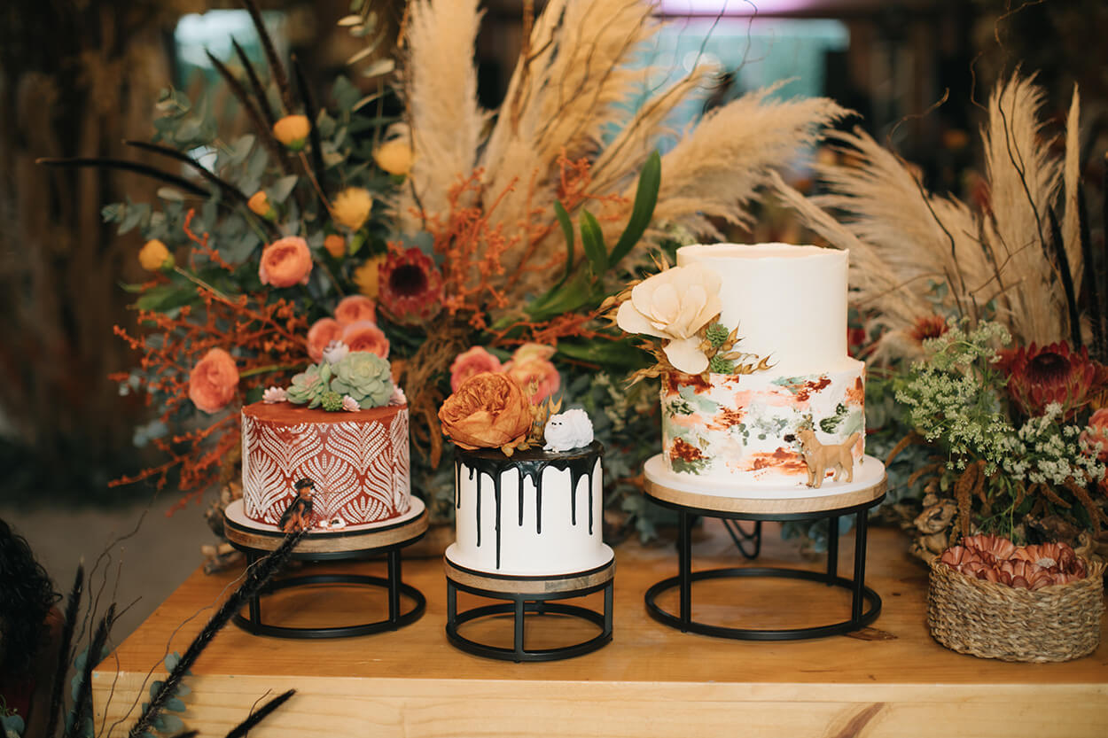 mesa de madeira com drip cake e flores laranja e vermelha em cima ao lado de bolo laranja com suculentas no topo e bolo branco de dois andares com pintura de flores e miniatura de cachorro