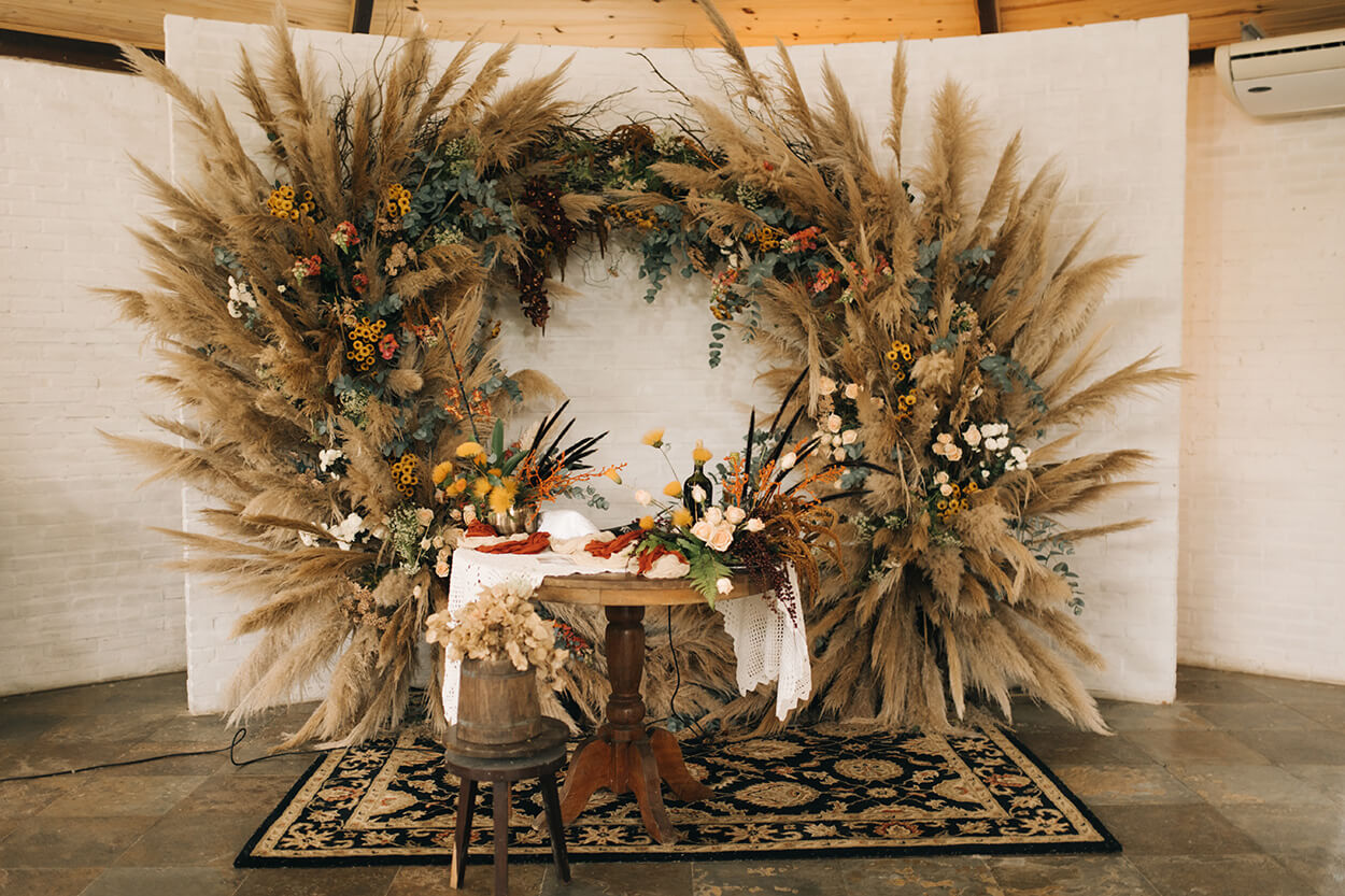 mesa de madeira redonda e ao fundo com arco com flores vermelhas amarelas e muito capim dos pampas
